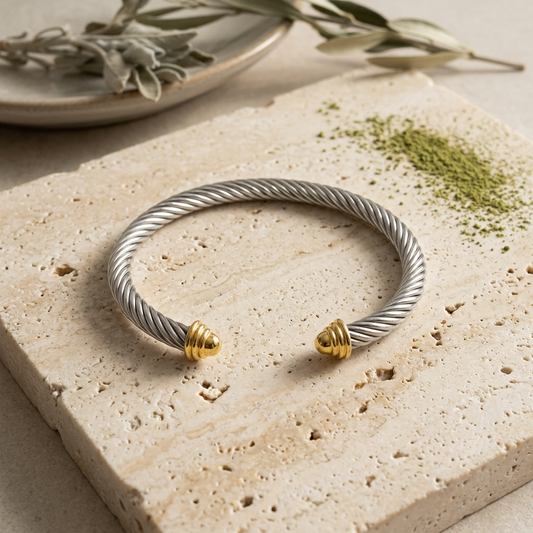 Pulsera plateada de cable trenzado con puntas doradas sobre piedra crema y un toque de polvo matcha.